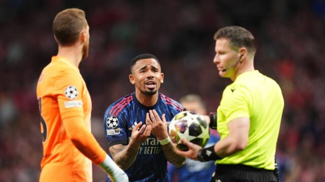 Zdá sa, že dvaja jednotlivci v športových uniformách, z ktorých jeden drží futbalovú loptu, diskutujú s rozhodcom zápasu v rušnom prostredí štadióna s divákmi viditeľnými v pozadí.