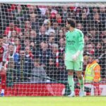 Dvaja jednotlivci oblečení v atletických uniformách sú postavení na futbalovom ihrisku, pričom jeden stojí za sieťou. Zdá sa, že pozorujú scénu zahŕňajúcu inú osobu, možno hráča, pred značným davom usadeným na tribúne. Celková atmosféra naznačuje intenzívny moment počas futbalového zápasu.