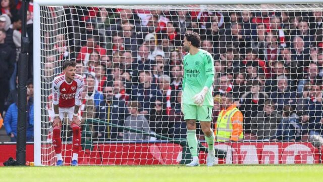 Dvaja jednotlivci oblečení v atletických uniformách sú postavení na futbalovom ihrisku, pričom jeden stojí za sieťou. Zdá sa, že pozorujú scénu zahŕňajúcu inú osobu, možno hráča, pred značným davom usadeným na tribúne. Celková atmosféra naznačuje intenzívny moment počas futbalového zápasu.