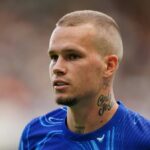 Muž s vyholenou hlavou a viditeľným tetovaním na krku má oblečený modrý športový dres. Stojí na ihrisku, pravdepodobne sa zúčastňuje súťažného športového podujatia, a zdá sa, že je zameraný na niečo v diaľke. Pozadie je mierne rozmazané, čo naznačuje pohyb alebo dav divákov.