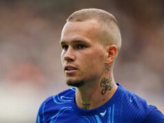 Hviezda Chelsea Mykhailo Mudryk „vydala štvorročný zákaz dopingu“ s Blues, ktorí sa chcú odvolať Muž s vyholenou hlavou a viditeľným tetovaním na krku má oblečený modrý športový dres. Stojí na ihrisku, pravdepodobne sa zúčastňuje súťažného športového podujatia, a zdá sa, že je zameraný na niečo v diaľke. Pozadie je mierne rozmazané, čo naznačuje pohyb alebo dav divákov.