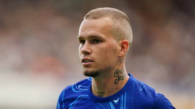 Muž s vyholenou hlavou a viditeľným tetovaním na krku má oblečený modrý športový dres. Stojí na ihrisku, pravdepodobne sa zúčastňuje súťažného športového podujatia, a zdá sa, že je zameraný na niečo v diaľke. Pozadie je mierne rozmazané, čo naznačuje pohyb alebo dav divákov.