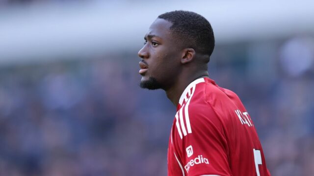 Športovec v červenom športovom drese s číslom 18 stojí na ihrisku, možno počas futbalového zápasu, s rozmazaným davom v pozadí.