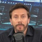 Jednotlivec s bradou, oblečený v tmavej košeli s dlhým rukávom, vyzerá ako zapojený do rozhovoru. Mikrofón je umiestnený pred osobou a na pozadí je rozmazané zobrazenie toho, čo vyzerá ako športové miesto.