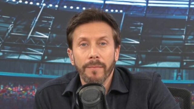 Jednotlivec s bradou, oblečený v tmavej košeli s dlhým rukávom, vyzerá ako zapojený do rozhovoru. Mikrofón je umiestnený pred osobou a na pozadí je rozmazané zobrazenie toho, čo vyzerá ako športové miesto.