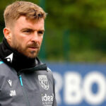 James Morrison v sivej teplákovej súprave West Bromwich Albion a čiernych nohaviciach na tréningu so slovom "BOSS" vidieť na rozmazanej reklamnej tabuli v diaľke za ním.