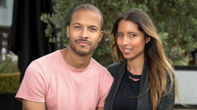 Obrázok zobrazuje dospelého muža a ženu, ktorí stoja blízko pri sebe, usmievajú sa a pózujú pre fotografiu vo vonkajšom prostredí. Sú obklopené zeleňou a kulisou, ktorá naznačuje prostredie záhrady alebo parku. Samec je oblečený vo svetloružovom tričku, zatiaľ čo samica má oblečené sako tmavej farby cez čierny top.