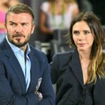 Obraz zobrazuje muža a ženu stojacich blízko, pravdepodobne na spoločenskej udalosti. Muž má na sebe okuliare a modrý kabát, žena má športové slnečné okuliare a biely top. V pozadí sú viditeľné rozmazané postavy, čo naznačuje preplnené prostredie.