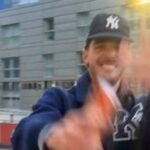 Jednotlivec oblečený v modrej bunde a baseballovej čiapke nadšene máva do kamery, zatiaľ čo stojí vo vonkajšom prostredí s modernou budovou viditeľnou v pozadí.