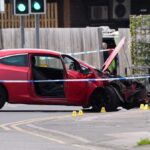 Scéna zobrazuje červené športové auto, ktoré bolo účastníkom nehody. Predná časť vozidla je značne poškodená a kapota je otvorená. Cesta je ohradená policajnou páskou, na mieste je prítomný policajt, ​​ktorý stojí neďaleko vozidla. V pozadí vidno semafor a drevený plot.