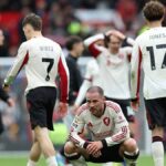 Skupina futbalistov na ihrisku, pričom jeden hráč kľačí a zdá sa, že sa rozpráva s druhým. Hráči majú oblečené biele a čierne uniformy a na pozadí je rozmazaný dav divákov.