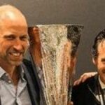 Traja muži oblečení v slávnostnom oblečení stoja tesne vedľa seba, pričom jeden drží veľkú trofej a ďalší dvaja ju prezentujú. Sú umiestnené na pozadí s výraznými oranžovými a šedými diagonálnymi pruhmi.