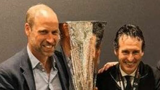 Traja muži oblečení v slávnostnom oblečení stoja tesne vedľa seba, pričom jeden drží veľkú trofej a ďalší dvaja ju prezentujú. Sú umiestnené na pozadí s výraznými oranžovými a šedými diagonálnymi pruhmi.