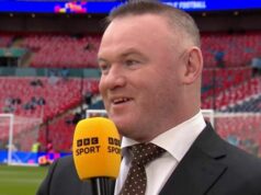Wayne Rooney hospitalizoval na natáčaní relácie BBC s legendou Man Utd, ktorej „hrozí operácia“ Muž oblečený vo formálnom obleku a kravate stojí na športovom ihrisku a drží mikrofón so žltým logom. V pozadí je prázdny štadión s červenými sedadlami a bránkou na opačnom konci.