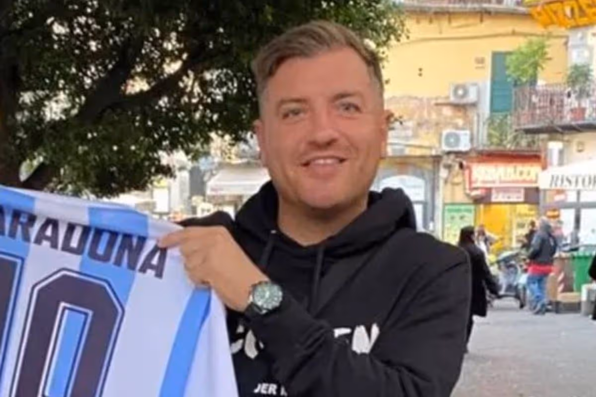 Jednotlivec oblečený v čiernej mikine s kapucňou zobrazuje dres s číslom 10 a stojí vo vonkajšom prostredí so stromom a rôznymi mestskými prvkami v pozadí.