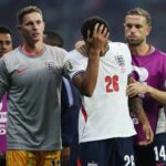 Futbalisti v tímových dresoch stoja na ihrisku. Jeden hráč sa drží rukou za hlavu, pravdepodobne v dôsledku faulu alebo zranenia,