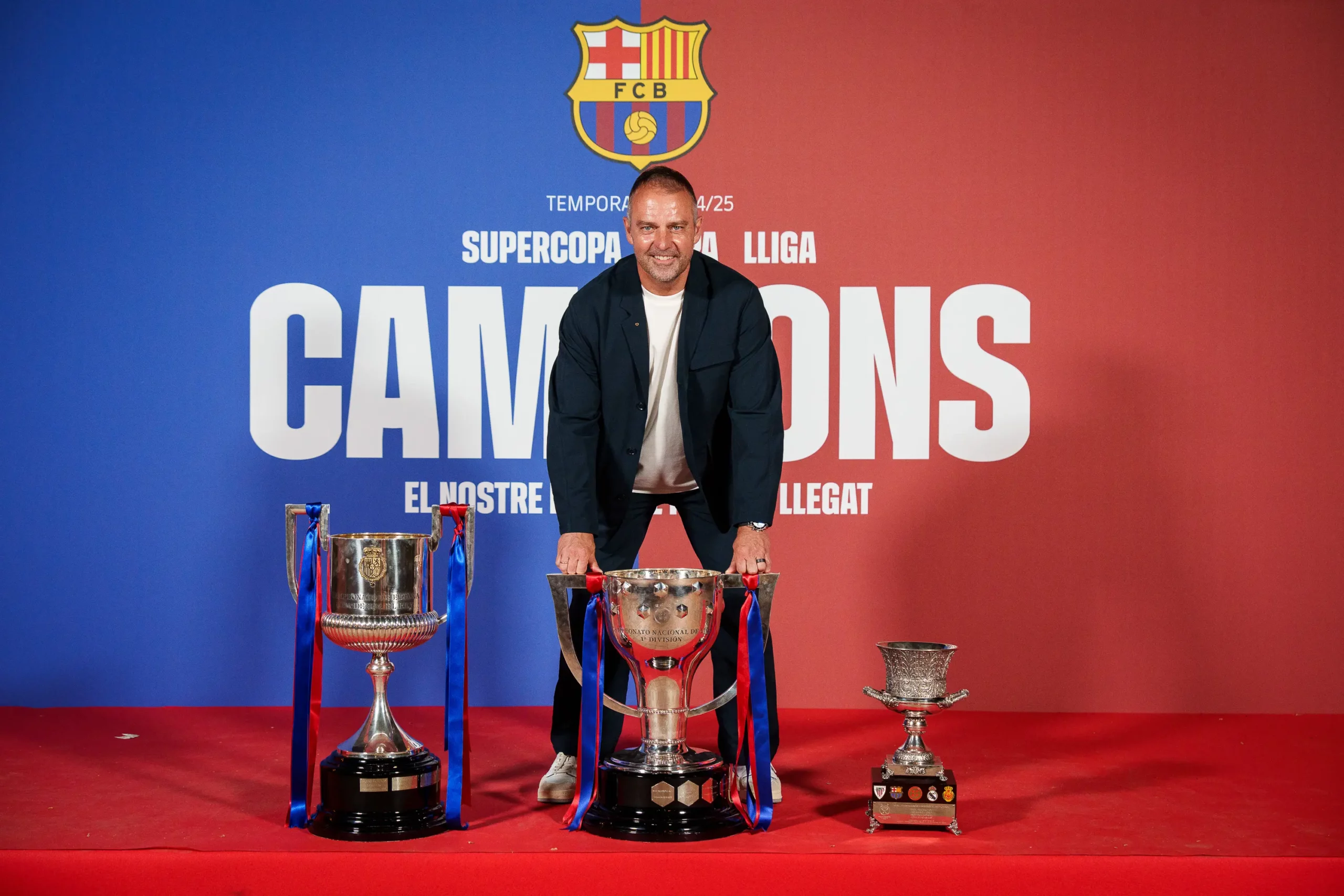 Manažér Barcelony Hansi Flick pózuje s trofejami na červenom koberci.