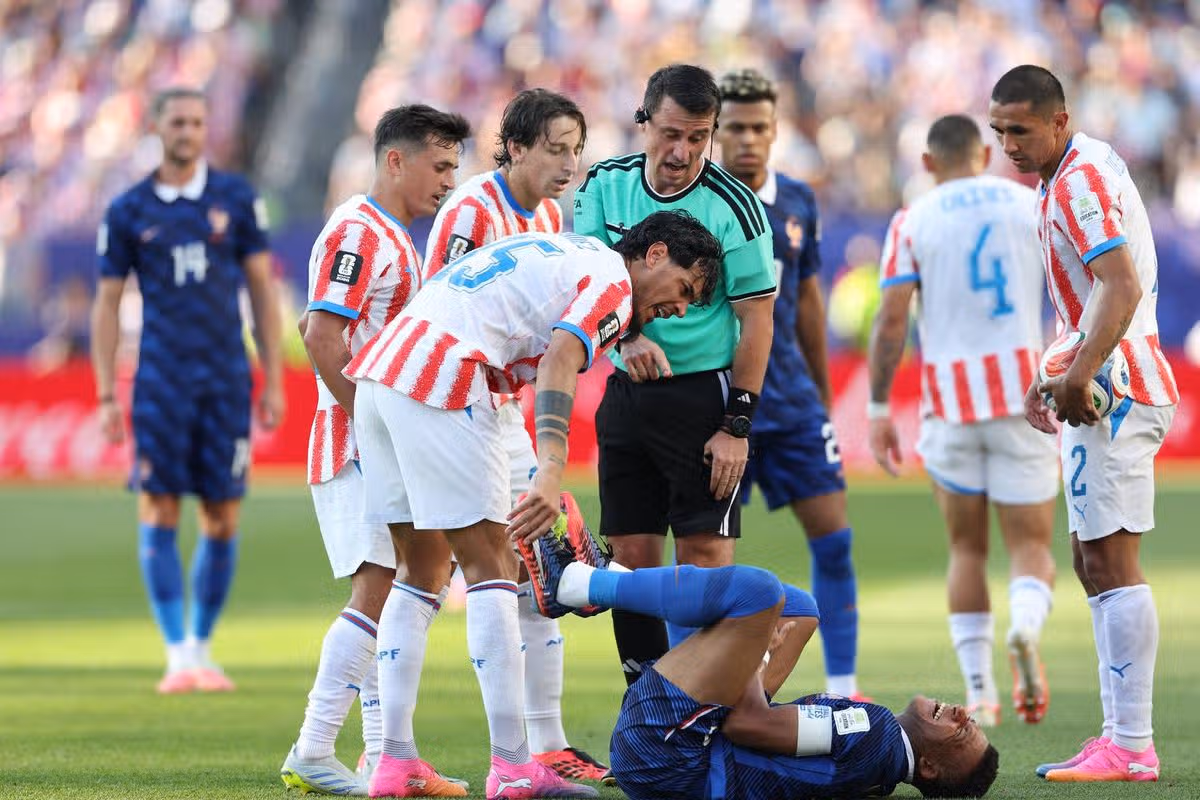 Skupina futbalistov na ihrisku, z ktorých jeden hráč leží na zemi a dostáva pozornosť od zdravotníckeho personálu, zatiaľ čo ostatní hráči a funkcionári to pozorujú.